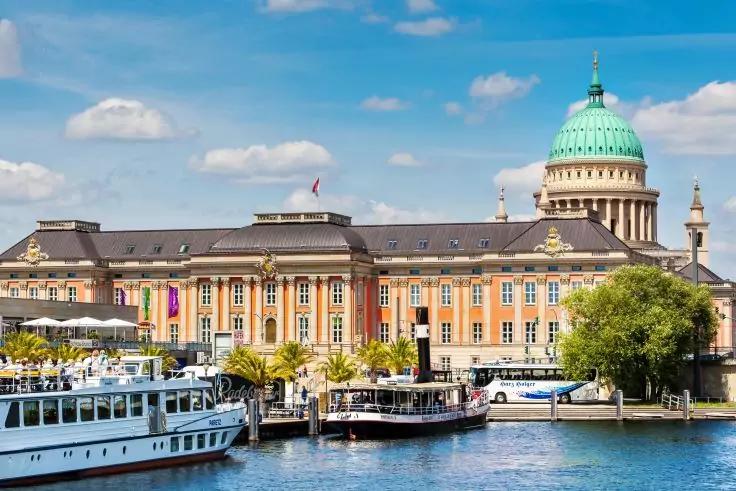 Gro&szlig;es gelblich-beiges Geb&auml;ude mit gr&uuml;ner Kuppel am Ufer, mehrere wei&szlig;e Ausflugsschiffe im Wasser, B&auml;ume und blauer Himmel mit Wolken