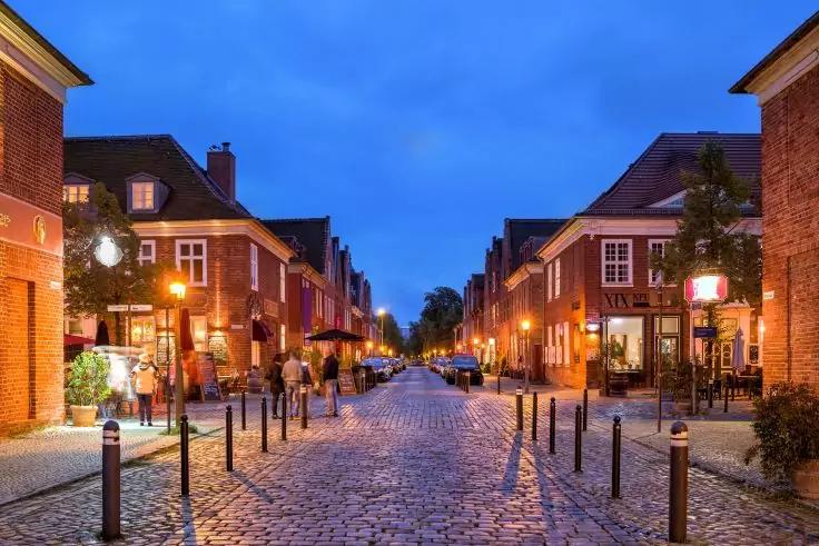 Pflasterstra&szlig;e bei D&auml;mmerung, von Backsteingeb&auml;uden ges&auml;umt, beleuchtete Laternen, Poller und Fu&szlig;g&auml;nger