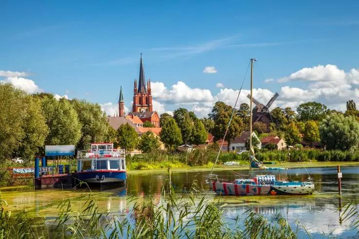 Flussufer mit Booten im Vordergrund, Schilf, B&auml;ume, Wohnh&auml;user, eine rote Backsteinkirche mit hohen T&uuml;rmen und eine Windm&uuml;hle unter blauem Himmel mit Wolken
