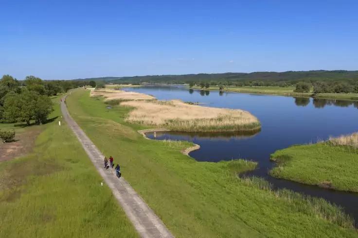 Flussufer mit Schilfinseln, unbefestigtem Weg entlang des Wassers, drei Personen auf dem Weg, blauer Himmel