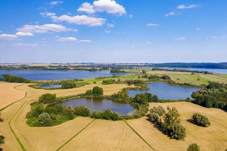 Luftaufnahme einer Landschaft mit mehreren kleinen Seen, Feldern und Baumgruppen, im Hintergrund ein gro&szlig;er See, blauer Himmel mit Wolken