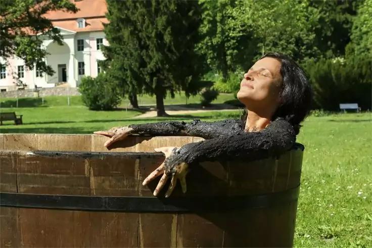 Person mit geschlossenen Augen und nach oben geneigtem Kopf lehnt mit Armen am Rand eines h&ouml;lzernen Bottichs auf einer Rasenfl&auml;che, im Hintergrund B&auml;ume und ein Geb&auml;ude