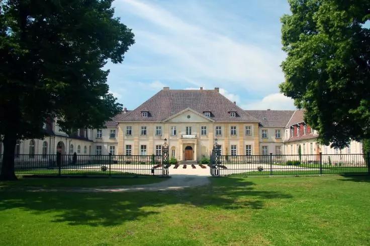 Gro&szlig;es hellgelbes Geb&auml;ude mit mittigem Eingang und symmetrischen Fl&uuml;geln, eiserner Zaun vorn, gr&uuml;ner Rasen, B&auml;ume links und rechts, blauer Himmel