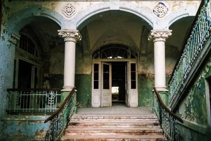 Breite Treppe mit gebogenen Handl&auml;ufen, f&uuml;hrt zu offenen Doppelt&uuml;ren zwischen S&auml;ulen; gr&uuml;nlich abgebl&auml;tterte W&auml;nde und dekorative Eisenbalustraden