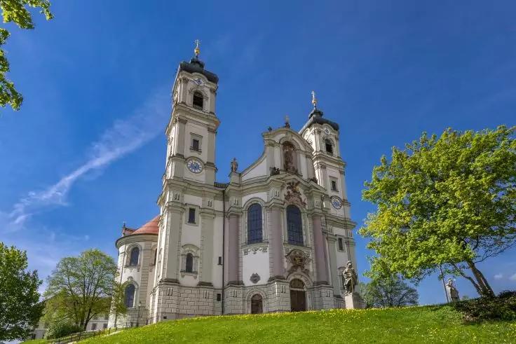 Hellbeiges Gebäude mit zwei hohen Türmen, Kreuzen auf den Türmchen, großen Bogenfenstern und verzierter Fassade, Steintreppe zum Haupteingang, grüner Rasen und Bäume, blauer Himmel mit wenigen Wolken
