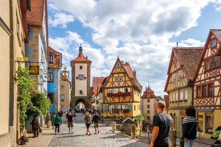 Kopfsteinpflasterstraße mit Fachwerkhäusern, einem Torturm mit Uhr im Hintergrund, mehreren Personen und blauem Himmel mit Wolken