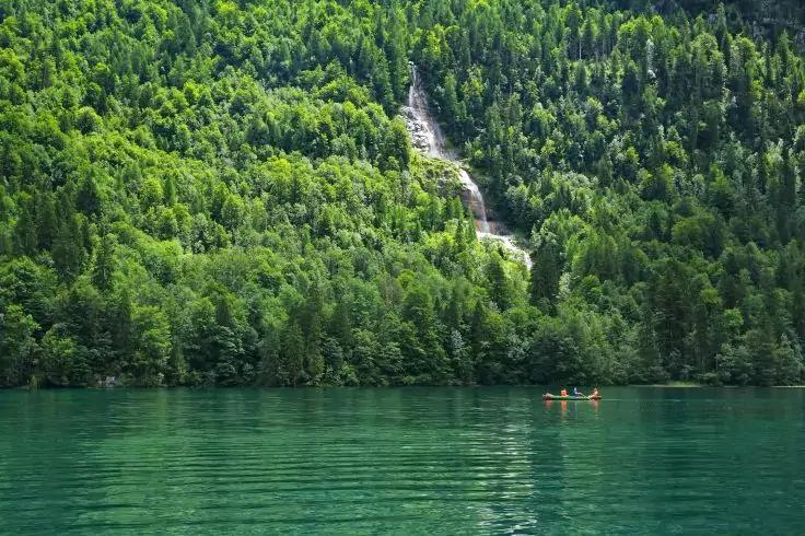 See vor dicht bewaldeter Bergflanke mit Wasserfall, auf dem See zwei Kajaks mit Personen
