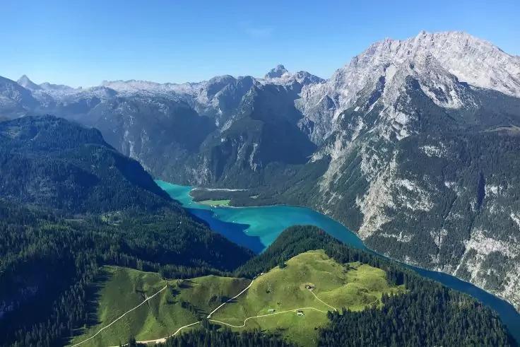 Grüne Bergwiese mit Wegen und einigen Hütten im Vordergrund, dahinter ein türkisfarbener See und steile felsige Berge unter klarem blauem Himmel