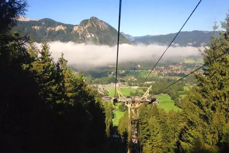 Seilbahnseile und Stütze über bewaldetem Abhang, Tal mit Wiesen und Ort, Nebelbank, Berge im Hintergrund und blauer Himmel