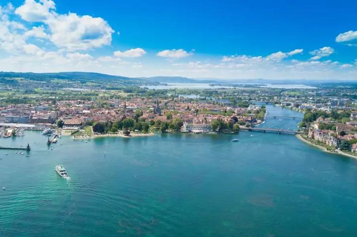 Luftaufnahme einer Uferstadt auf einer Halbinsel mit Gebäuden, Brücken und Booten auf blauem Wasser, grünen Hügeln und wolkigem Himmel