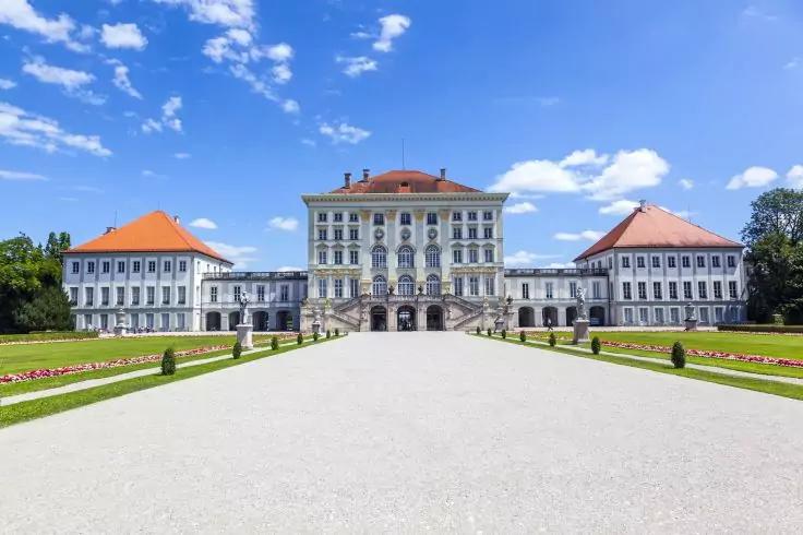 Großes symmetrisches Gebäude mit mittlerem Haupttrakt und zwei seitlichen Flügeln, roten Dächern, breitem Kiesweg im Vordergrund, grünen Rasenflächen und Blumenbeeten, blauem Himmel mit Wolken
