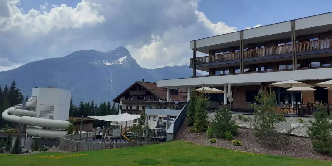 Mehrst&ouml;ckiges Geb&auml;ude mit Balkonen, Terrasse mit Sonnenschirmen, geschwungene wei&szlig;e Rutsche, Holzhaus, Rasen, Nadelb&auml;ume und Berg mit Schneeflecken unter bew&ouml;lktem Himmel
