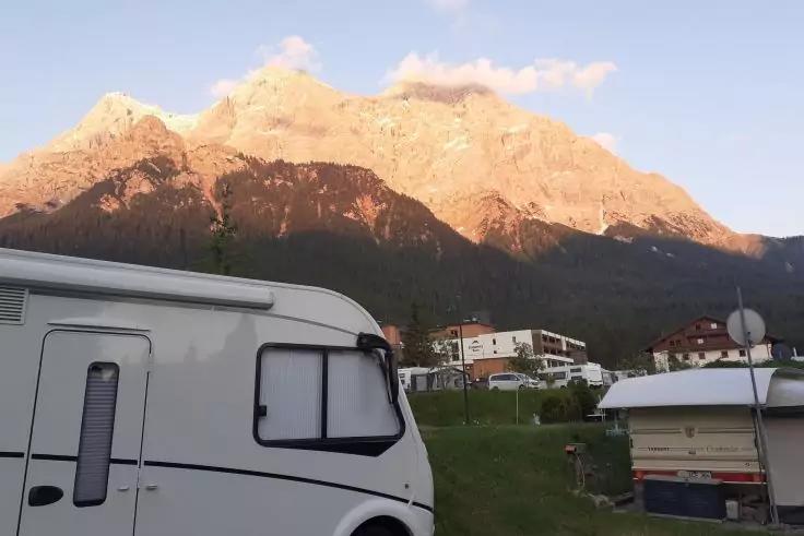 Weißes Wohnmobil links im Vordergrund, rechts kleiner Wohnwagen, Häuser auf grasiger Fläche in der Bildmitte, Berge im Hintergrund mit warmem Licht, blauer Himmel mit wenigen Wolken
