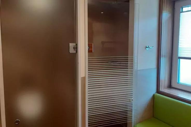 Innenraum mit braunen Holzpaneelen, links eine braune Tür, in der Mitte eine Glaswand mit horizontalen Milchglasstreifen, rechts ein Fenster mit weißen Jalousien und ein grünes Sitzkissen