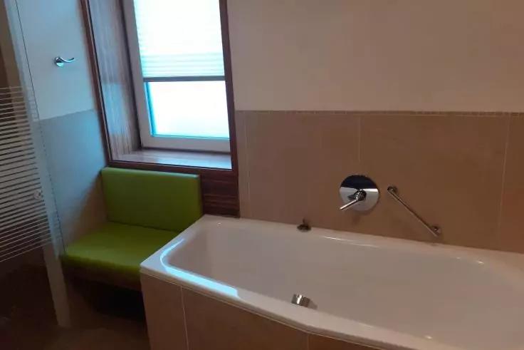 Weiße Badewanne mit verchromtem Wasserhahn, beige geflieste Wand rechts, Fenster mit weißer Jalousie und grüner gepolsterter Sitzfläche links