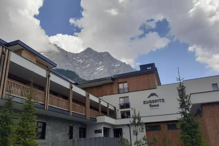 Gebäudefront mit Holzbalustraden und Schriftzug 'ZUGSPITZ', Nadelbäume, schneebedeckter Berg im Hintergrund unter bewölktem Himmel