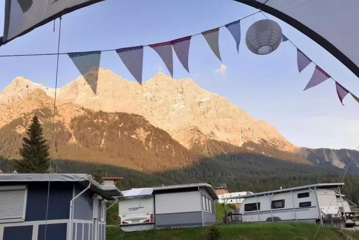 Wimpelgirlande und Papierlaterne über mehreren Wohnwagen und Stellplätzen mit Gras, im Hintergrund sonnenbeschienene Berge