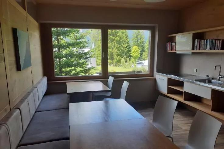 Innenraum mit Holzpanelen und -möbeln: zwei Tische, mehrere Stühle, gepolsterte Sitzbank entlang der linken Wand, großes rechteckiges Fenster mit Blick auf Bäume, offene Regale mit Büchern und Küchenzeile mit Spüle und Schränken