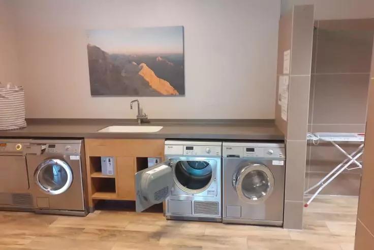 Drei silberne Frontlader unter einer Arbeitsplatte, mittlerer mit geöffneter Tür; Spüle mit Armatur darüber, Bild an der Wand, Wäschekorb links und Bügelbrett rechts