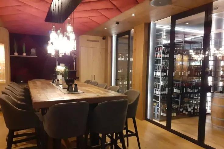Langer Holztisch mit gepolsterten Stühlen, Kronleuchter darüber, Glastür mit sichtbaren Weinregalen und Flaschen, Holzboden und hölzernes Fass