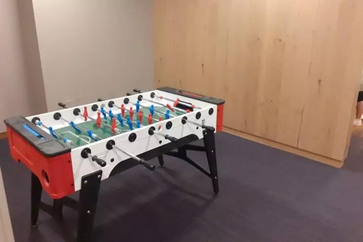 Tischkicker mit roten und blauen Spielfiguren, weiß-rotem Gehäuse und schwarzen Beinen auf grauem Teppich vor hellem Holzpaneel