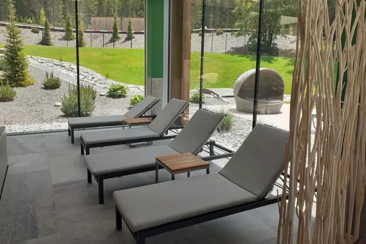 Vier graue Liegestühle mit kleinem Holztisch in einem verglasten Raum; draußen Rasen, Kiesflächen, Bepflanzung und eine runde Steinskulptur