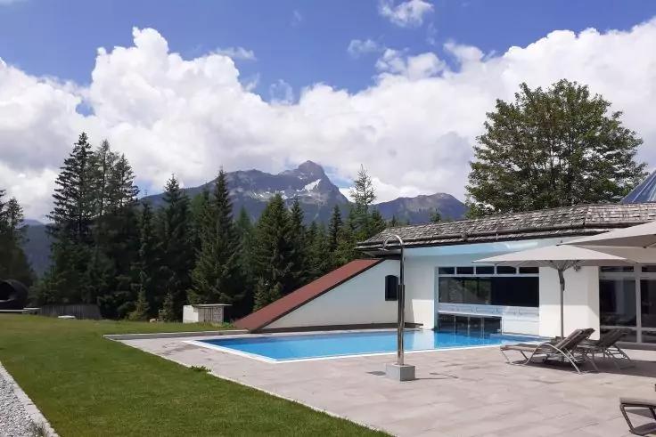 Außenpool neben modernem Gebäude mit Glasfront, Liegestühle und Sonnenschirm auf der Terrasse, Rasen, Nadelbäume, Berge und Wolken am blauen Himmel