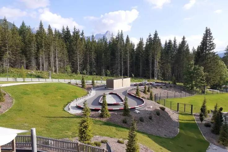 Freifläche mit runden Wasserbecken, Rasenflächen, kleinen Nadelbäumen, Holzzaun im Vordergrund, Waldhang, Berge und teils bewölkter Himmel