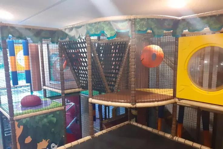 Indoor-Spielanlage mit gepolsterten Plattformen, Netzwänden, hängendem orangefarbenem Ball und gelbem Panel mit rundem Fenster