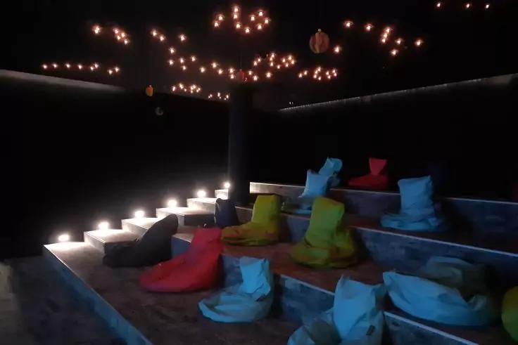 Dunkler Raum mit stufenförmigen Sitzebenen, bunten Sitzsäcken auf runden Podesten, beleuchteten Treppenstufen links und vielen kleinen hängenden Lichtern an der Decke