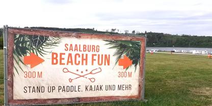 Gro&szlig;es Schild mit Text 'SAALBURG BEACH FUN' und darunter 'STAND UP PADDLE, KAJAK UND MEHR', seitliche Pfeile mit '300 m', Palmblatt-Grafiken und gekreuzte Paddel; im Hintergrund Wiese, Wasser mit Booten und bew&ouml;lkter Himmel