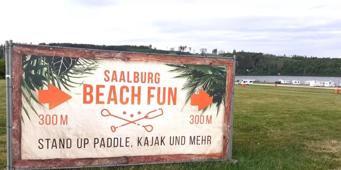 Gro&szlig;es Schild mit Text 'SAALBURG BEACH FUN' und darunter 'STAND UP PADDLE, KAJAK UND MEHR', seitliche Pfeile mit '300 m', Palmblatt-Grafiken und gekreuzte Paddel; im Hintergrund Wiese, Wasser mit Booten und bew&ouml;lkter Himmel