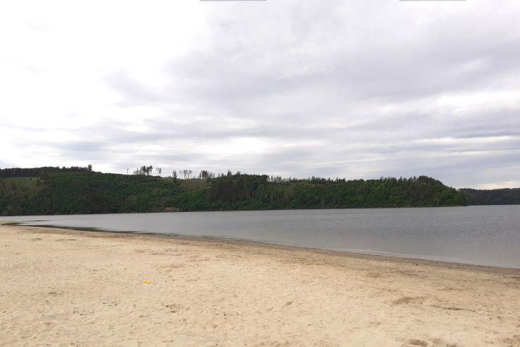 Sandstrand im Vordergrund, Wasserfl&auml;che, bewaldetes Ufer und bew&ouml;lkter Himmel