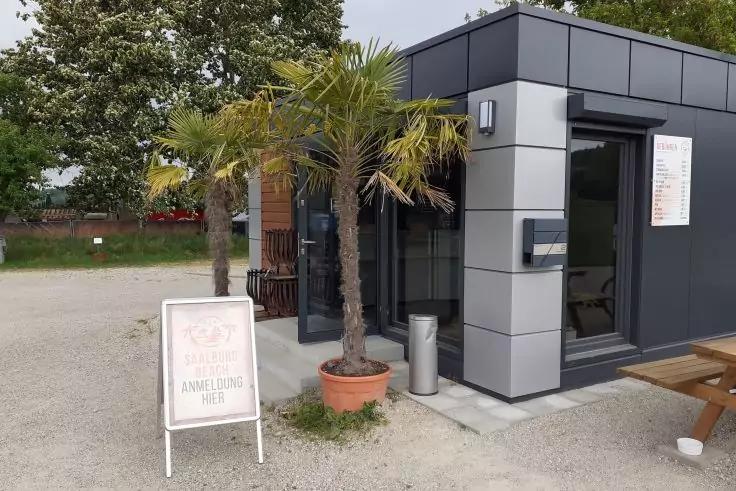 Kleines rechteckiges Geb&auml;ude mit Glasfront und grauen Metallpaneelen; davor zwei Palmen in Terrakottat&ouml;pfen, Kiesboden, eine Aufstelltafel links, Picknickbank rechts, runder Metallbeh&auml;lter neben dem Eingang, B&auml;ume und Rasen im Hintergrund