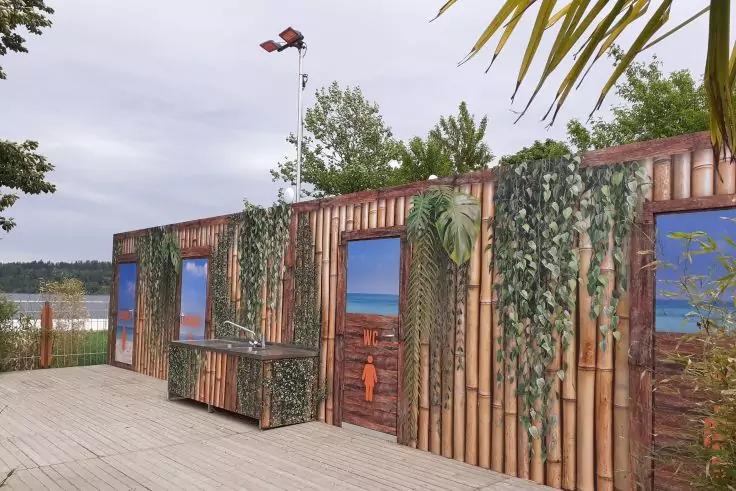 Bambusverkleidete Fassadenwand mit drei gemalten Fensterbildern des Meeres, h&auml;ngenden gr&uuml;nen Ranken, kleinem Ausschank/Kiosk, Holzdeck, Laternenmast, Wasser und B&auml;ume im Hintergrund, Palmenbl&auml;tter oben rechts, bew&ouml;lkter Himmel