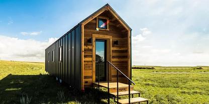 Kleines Holzhaus mit Glast&uuml;r, Treppe, seitlichen Fenstern und dreieckigem Fenster mit bunten Scheiben &uuml;ber der T&uuml;r auf einer gr&uuml;nen Wiese unter blauem Himmel mit Wolken
