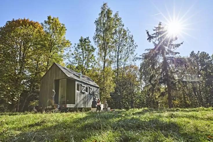 Kleines Holzhaus auf einer grasbewachsenen Lichtung, zwei Personen sitzen vor dem Haus, umgeben von B&auml;umen, Sonne mit Strahlen am Himmel