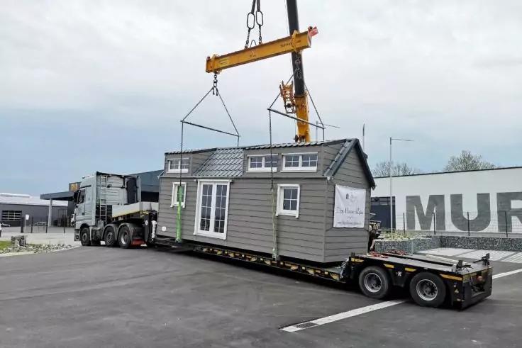 Graues kleines Hausmodul mit Fenstern und T&uuml;r, an Hebegurten unter einem Kran h&auml;ngend auf einem Tieflader vor einem Lkw; Asphaltfl&auml;che und Geb&auml;ude im Hintergrund