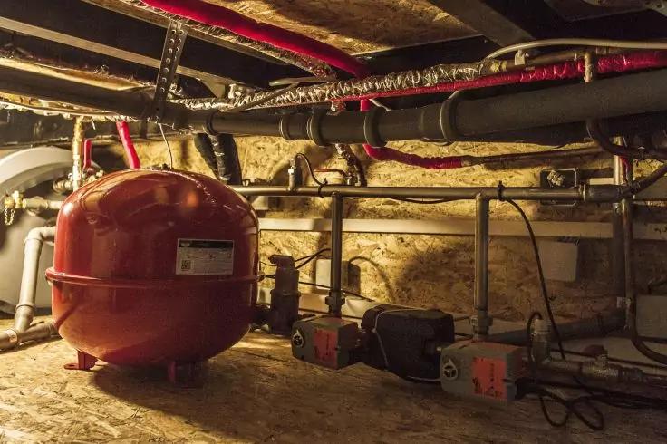 Roter runder Beh&auml;lter vor einer Steinwand, Metallrohre und isolierte Leitungen an der Decke, zwei Pumpen auf dem Boden