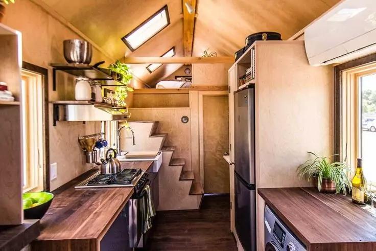 Schmale K&uuml;che in einem Dachraum mit Holzverkleidung, zwei Dachfenstern, Arbeitsfl&auml;che mit Sp&uuml;le und Herd, K&uuml;hlschrank, Waschmaschine, offene Regale mit Geschirr und Pflanzen, Treppe zu einer erh&ouml;hten Plattform, Wandklimaanlage, Fenster mit Topfpflanz