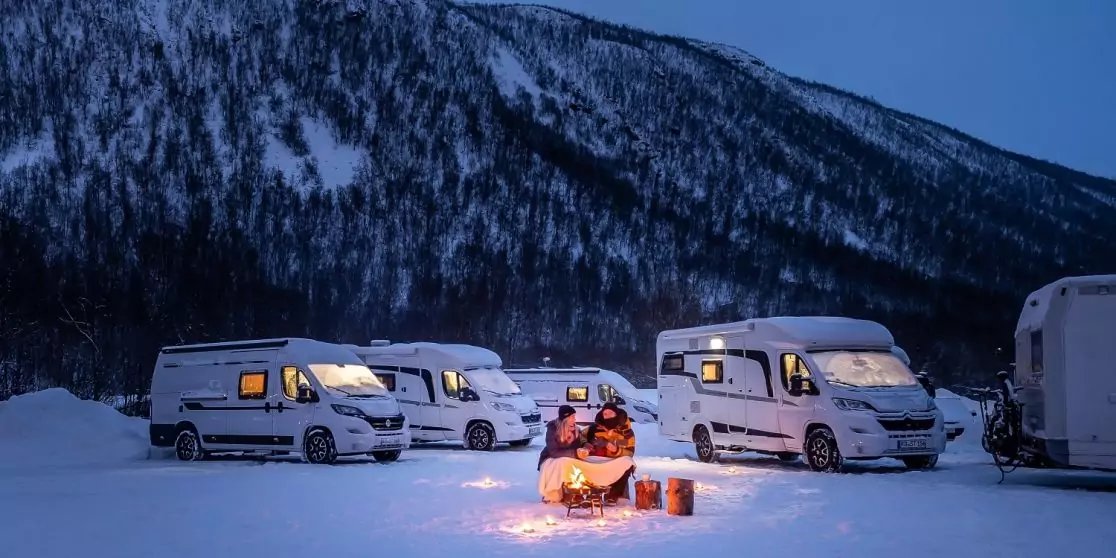 Mehrere wei&szlig;e Wohnmobile stehen bei D&auml;mmerung im Schnee vor einem schneebedeckten Berg; zwei Personen sitzen an einem kleinen Feuer neben einem Schneemobil