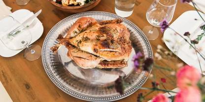 Ganzes H&auml;hnchen mit brauner Haut auf einer silbernen Servierplatte auf einem Holztisch; Teller, Gl&auml;ser und eine Sch&uuml;ssel im Hintergrund; unscharfe Blumen im Vordergrund