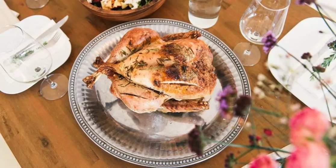 Ganzes H&auml;hnchen mit brauner Haut auf einer silbernen Servierplatte auf einem Holztisch; Teller, Gl&auml;ser und eine Sch&uuml;ssel im Hintergrund; unscharfe Blumen im Vordergrund
