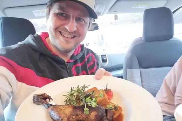 Lächelnder Mann mit Baseballcap sitzt im Auto und hält einen Teller mit gebratener Keule, Kräutern und Gemüse; rechter Arm einer weiteren Person ist teilweise sichtbar