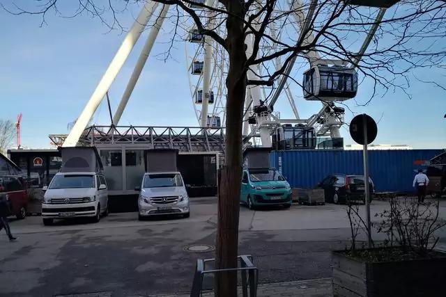 Parkfläche mit drei geparkten Fahrzeugen, mittiger Baumstamm im Vordergrund, dahinter ein großes Riesenrad mit Gondeln und blauen Containern