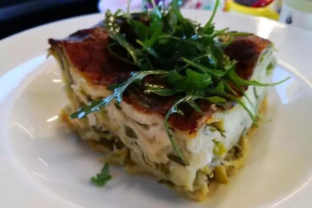 Ein Stück gebackenes Gericht mit goldbrauner Kruste und weißen Schichten, garniert mit grünen Blättern auf einem weißen Teller