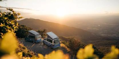 Zwei Wohnmobile auf einem H&uuml;gel mit Blick ins Tal, unscharfe gelbe Blumen im Vordergrund, Sonne am Horizont