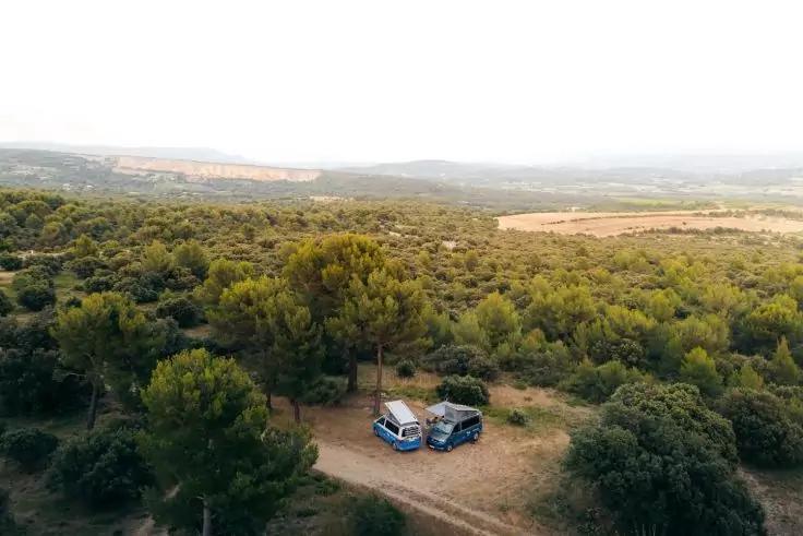 Luftaufnahme: zwei blaue Campervans in einer Lichtung, umgeben von B&auml;umen; H&uuml;gel im Hintergrund