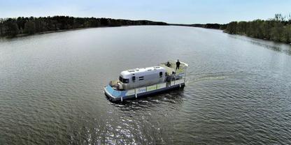Wei&szlig;es Hausboot auf ruhigem Fluss, eine Person auf dem Deck, baumbewachsene Ufer im Hintergrund