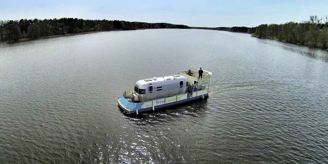 Wei&szlig;es Hausboot auf ruhigem Fluss, eine Person auf dem Deck, baumbewachsene Ufer im Hintergrund